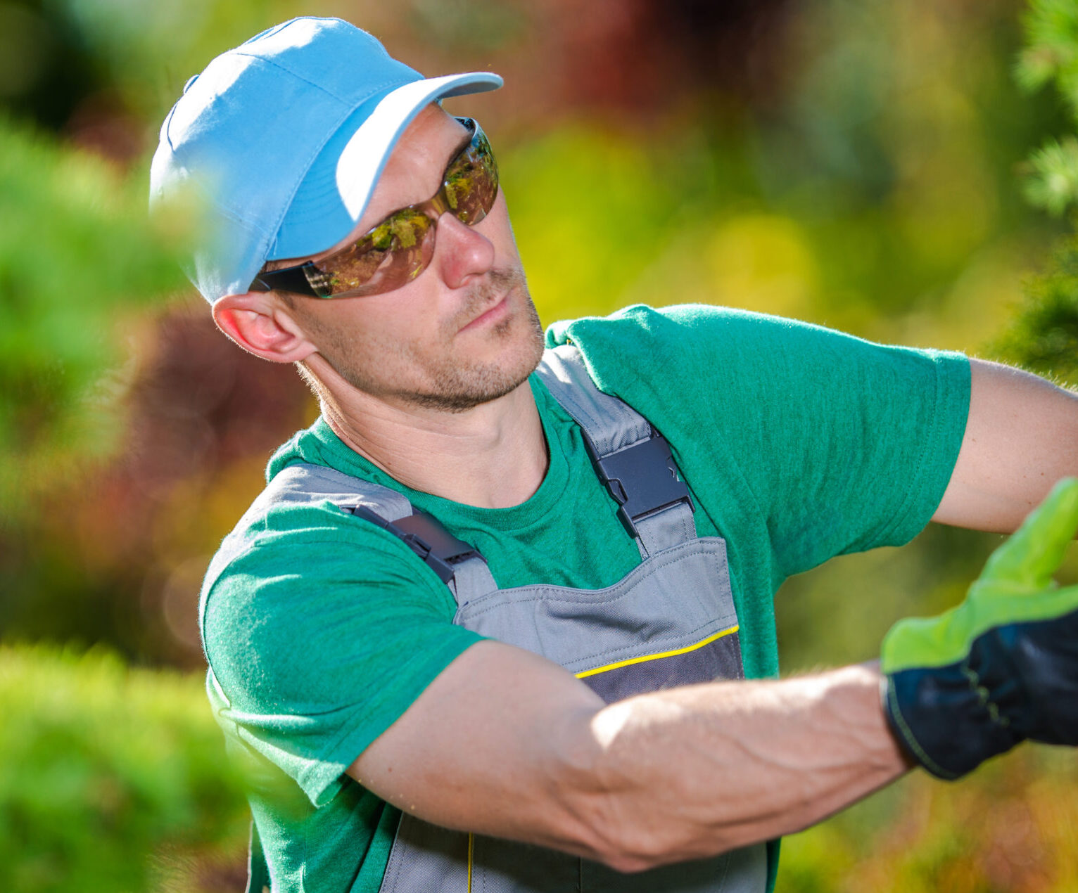 A gardener in protective gear carefully shapes plants in a vibrant garden under clear skies, showcasing detailed horticultural work.